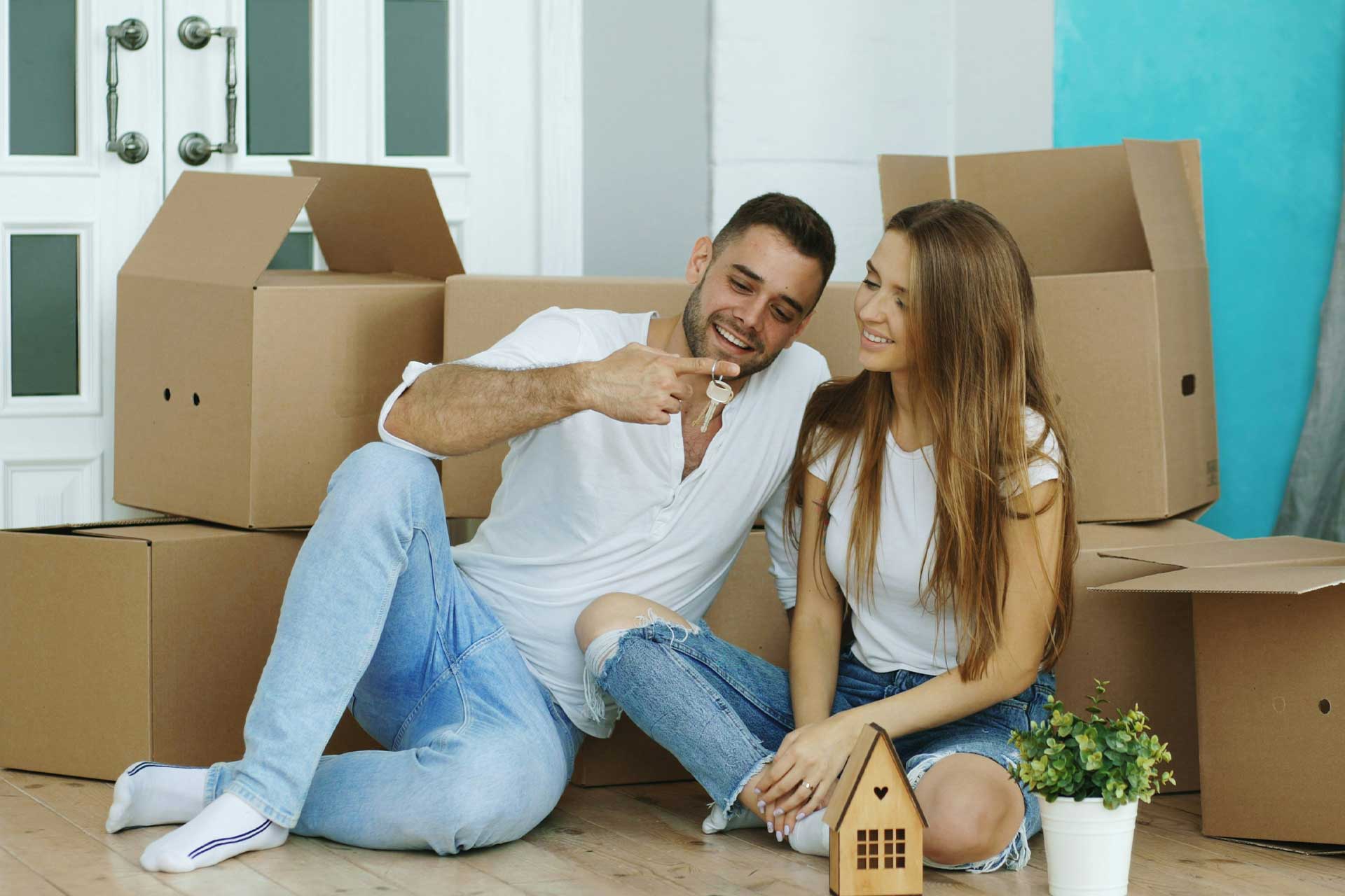 young couple sitting on ground of new home surrounding by moving boxes smiling holding new home keys